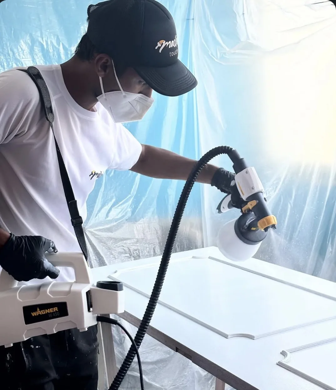 Painter in a mask spray-lacquering a door white inside a plastic-sheeted area