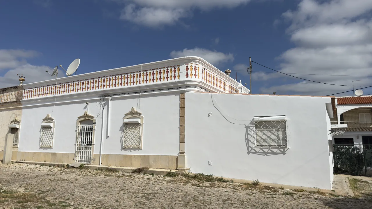 Traditional single-storey house painted white, with a two-tone balustraded parapet and barred windows, under a blue sky